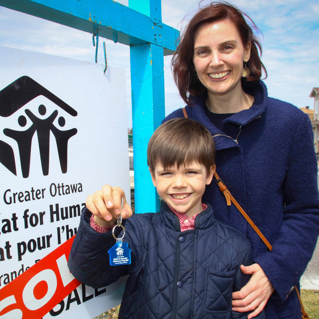 Habitat Partner Family mother and son standing in front of a sold sign. The child is holding up the keys to their Habitat home.