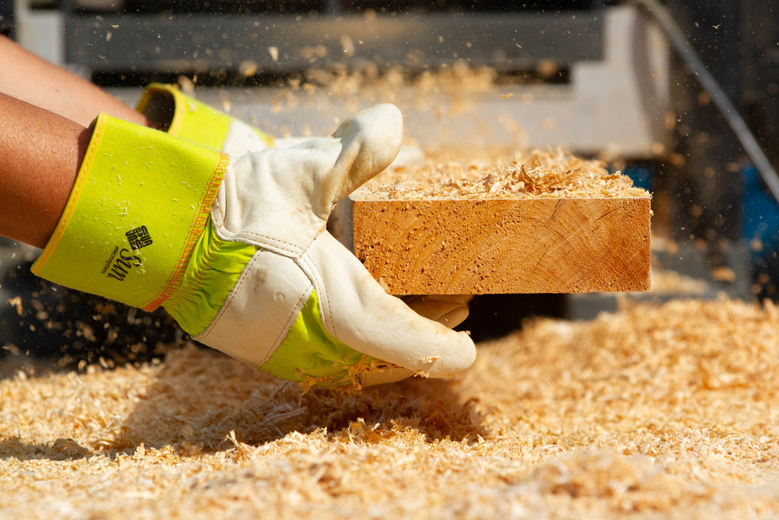 Hands wearing construction gloves putting a piece of lumber through an electric saw.