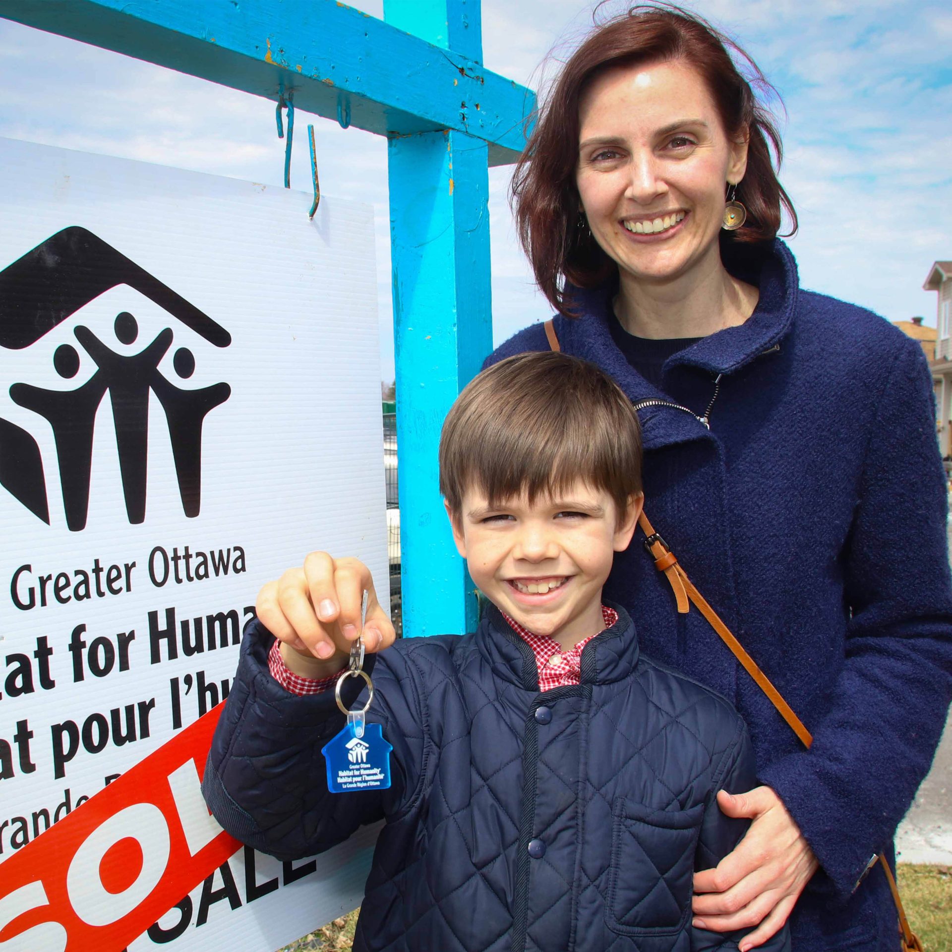 Mère et fils d'une famille partenaire Habitat debout devant une pancarte vendue. L'enfant tient les clés de leur maison Habitat.