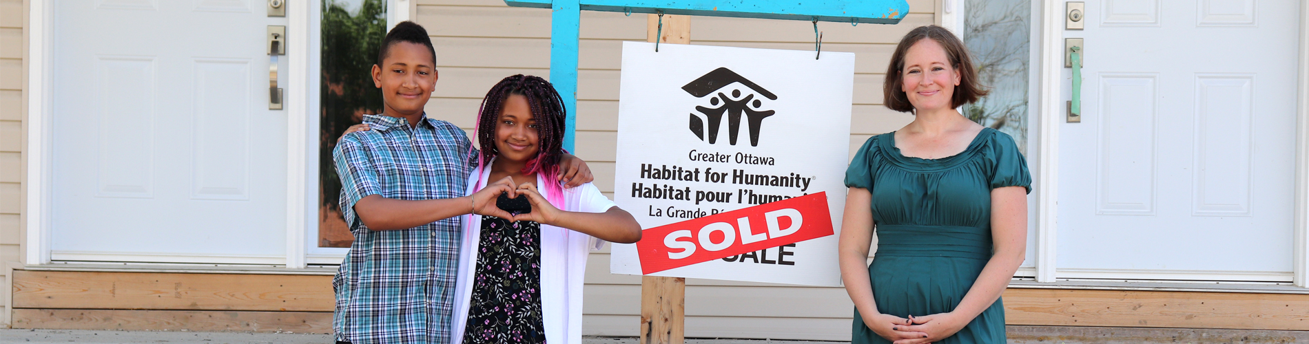 A Habitat Family Partner and her two children standing next to a "SOLD" sign in front of their Habitat home.