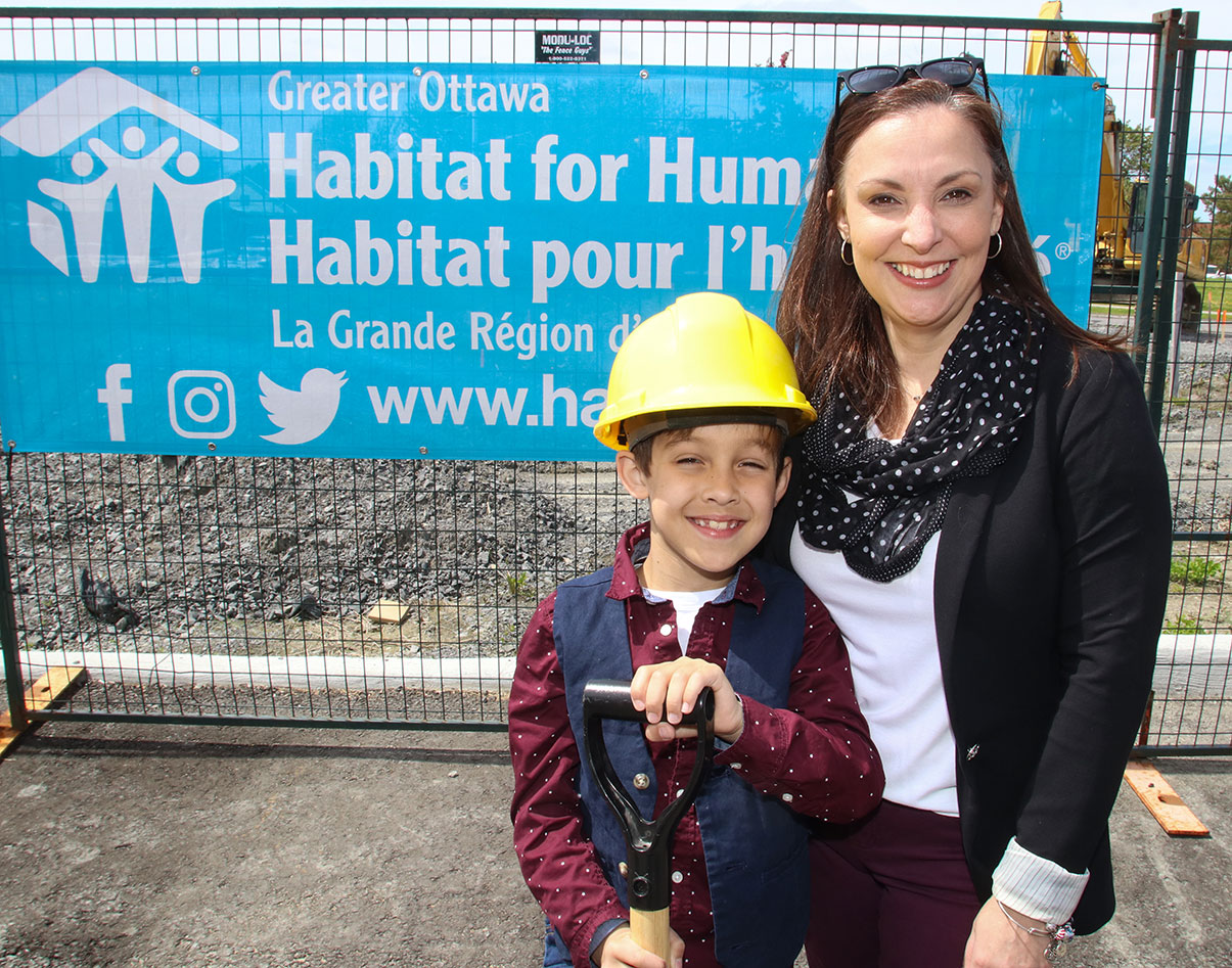 Habitat Partner Family Isabelle and her son wearing a construction hat and holding a shovel are standing outsiide in front of a Habitat Greater Ottawa banner during a Habitat Groundbreaking ceremony.