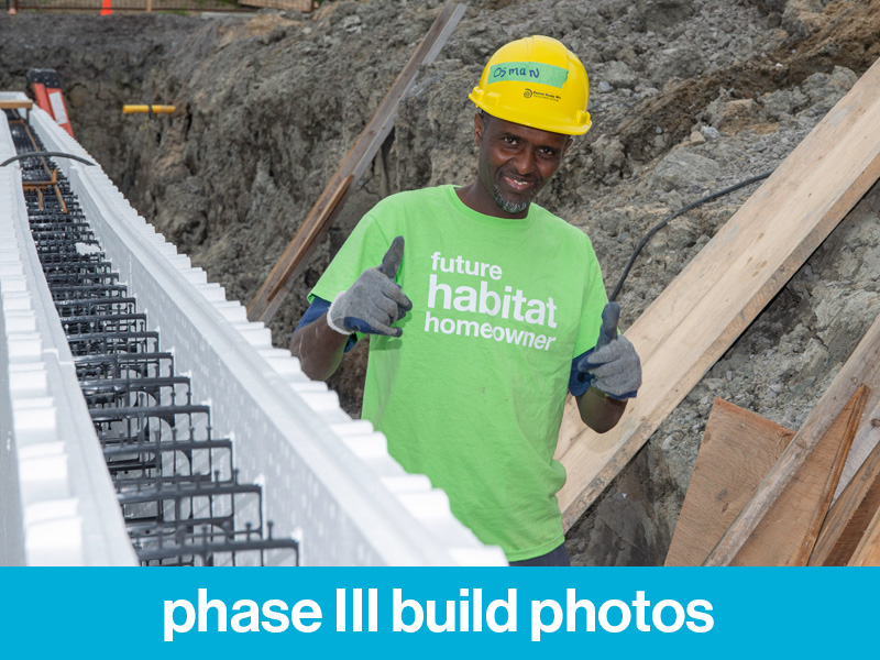 A Habitat Partner Family in a hard hat and a t-shirt that reads "future Habitat Homeowner" on a Habitat build site giving the thumbs up. The text at the bottom reads "phase III build photos"