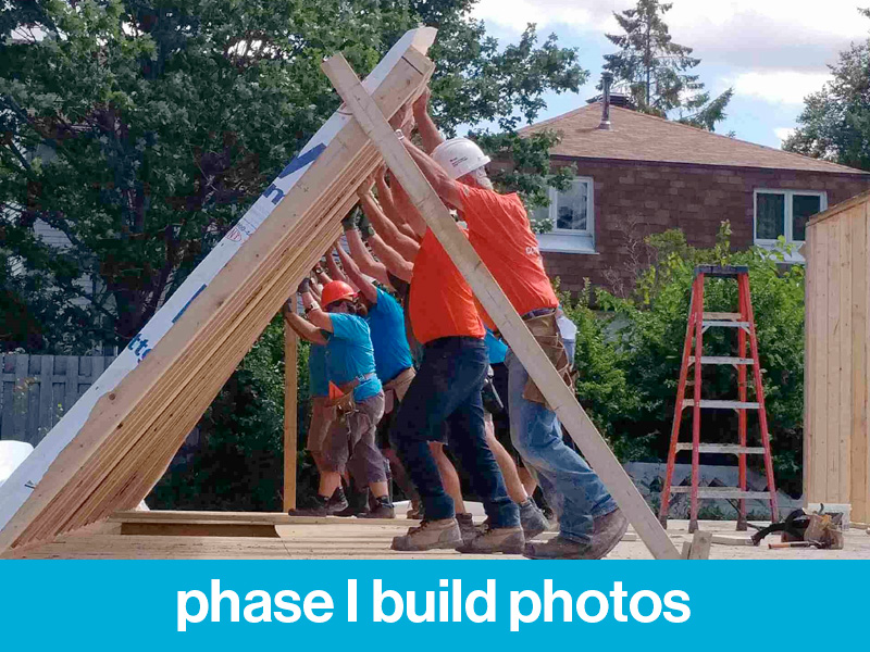 A group of Habitat build volunteers lifting up a wall. Text at bottom of image reads "phase I build photos".