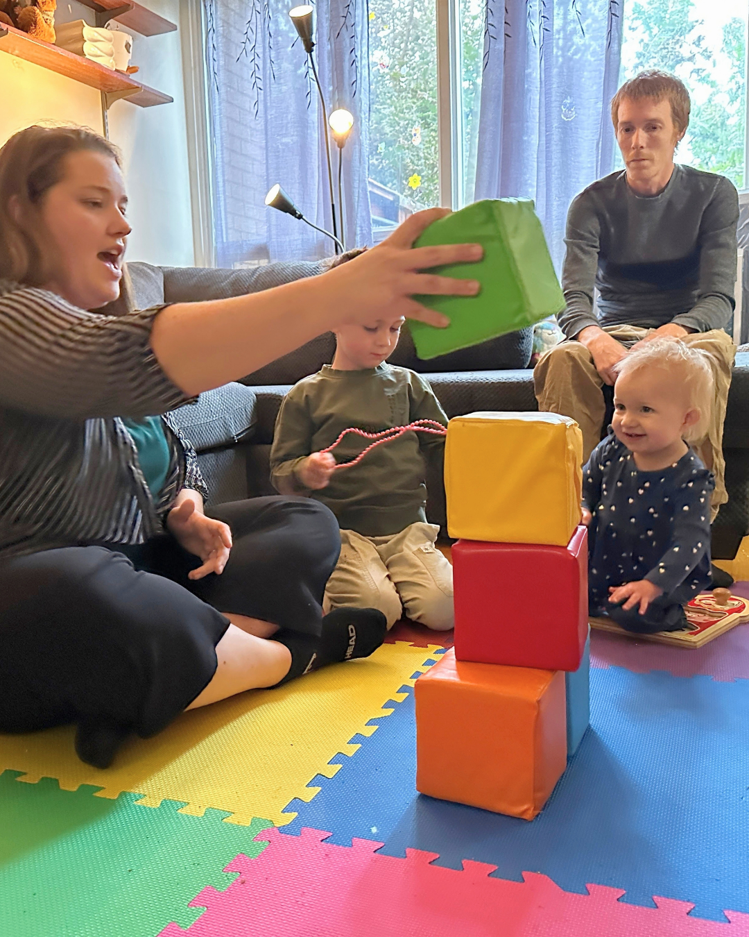 Kayla stacks blocks for Aubrey while Kale sits on the couch watching Dennis play with a bead necklace